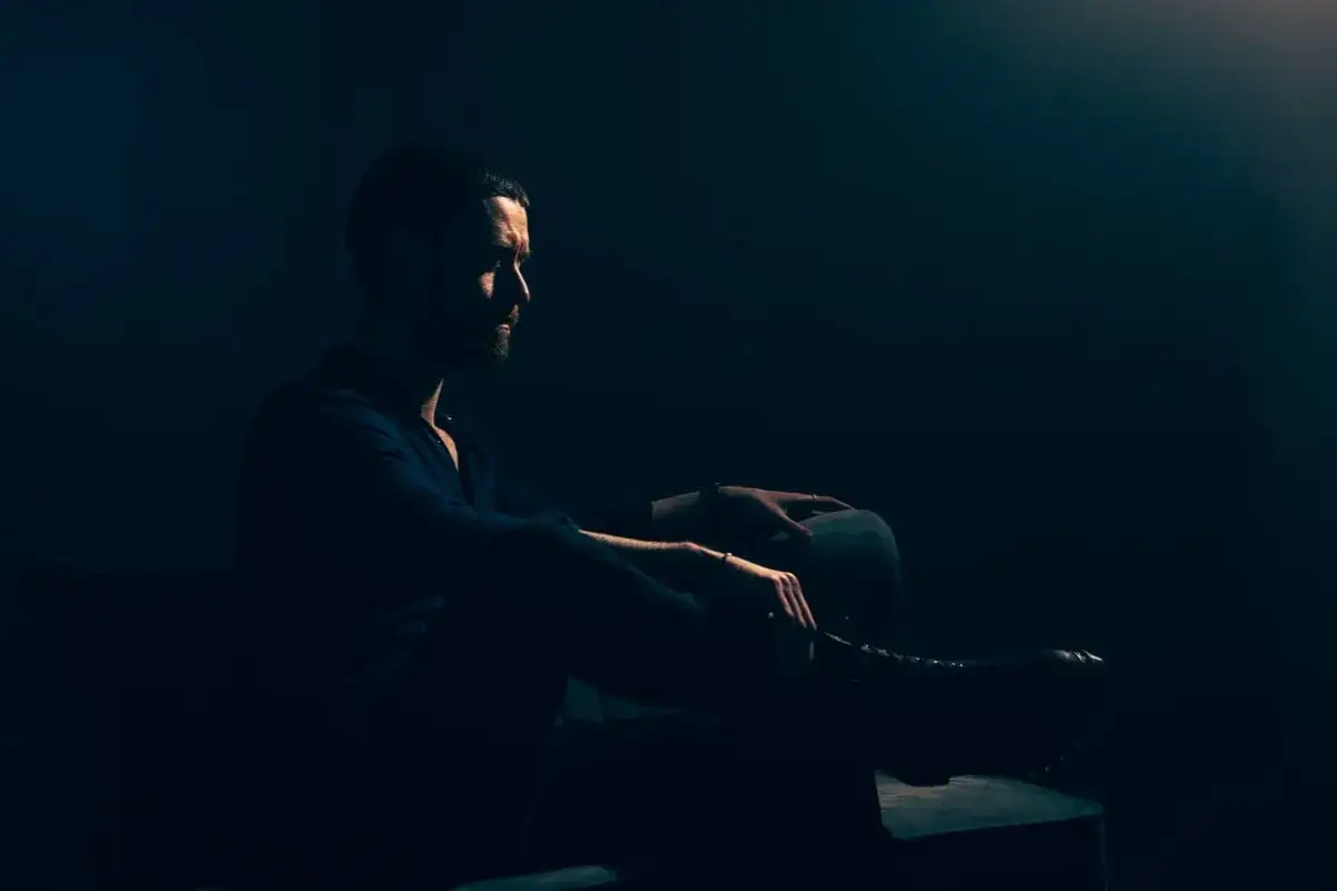 Uomo con barba seduto in penombra, illuminato da un fascio di luce laterale. Atmosfera da foto low key.