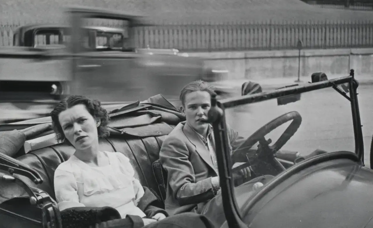 Coppia in auto, stile Walker Evans. Lei guarda pensierosa, lui al volante. Auto d'epoca in movimento.