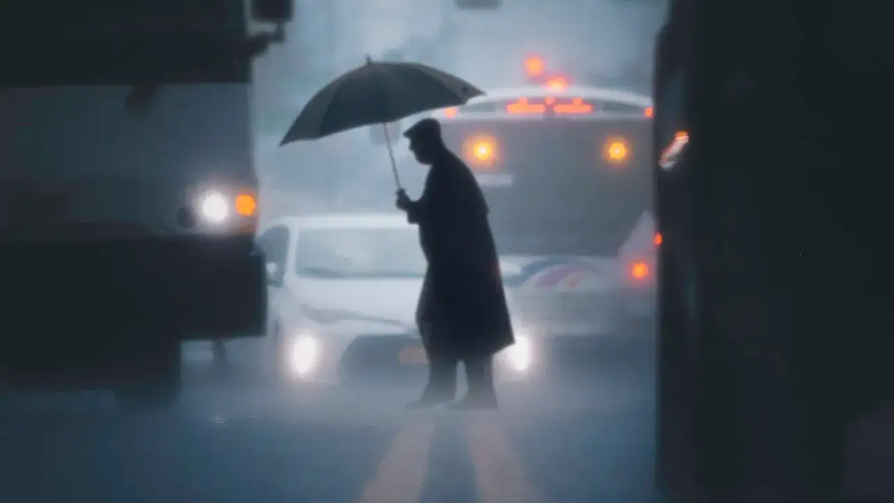 Uomo con ombrello attraversa la strada sotto la pioggia, circondato da luci sfocate di auto e bus. Una foto con la pioggia che crea un'atmosfera suggestiva.