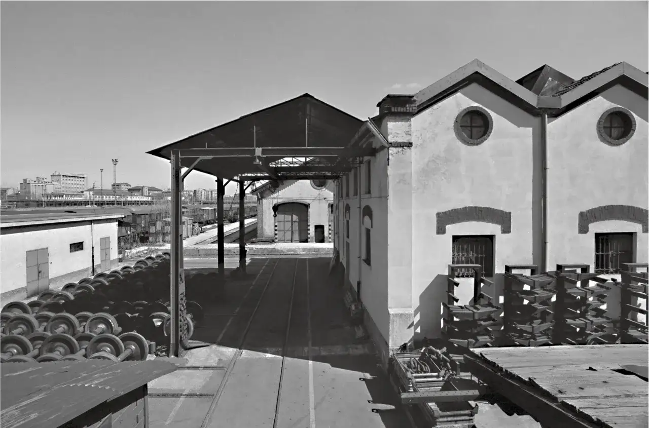 Gabriele Basilico foto: capannoni ferroviari, ruote di treno accatastate, binari e un paesaggio urbano sullo sfondo.