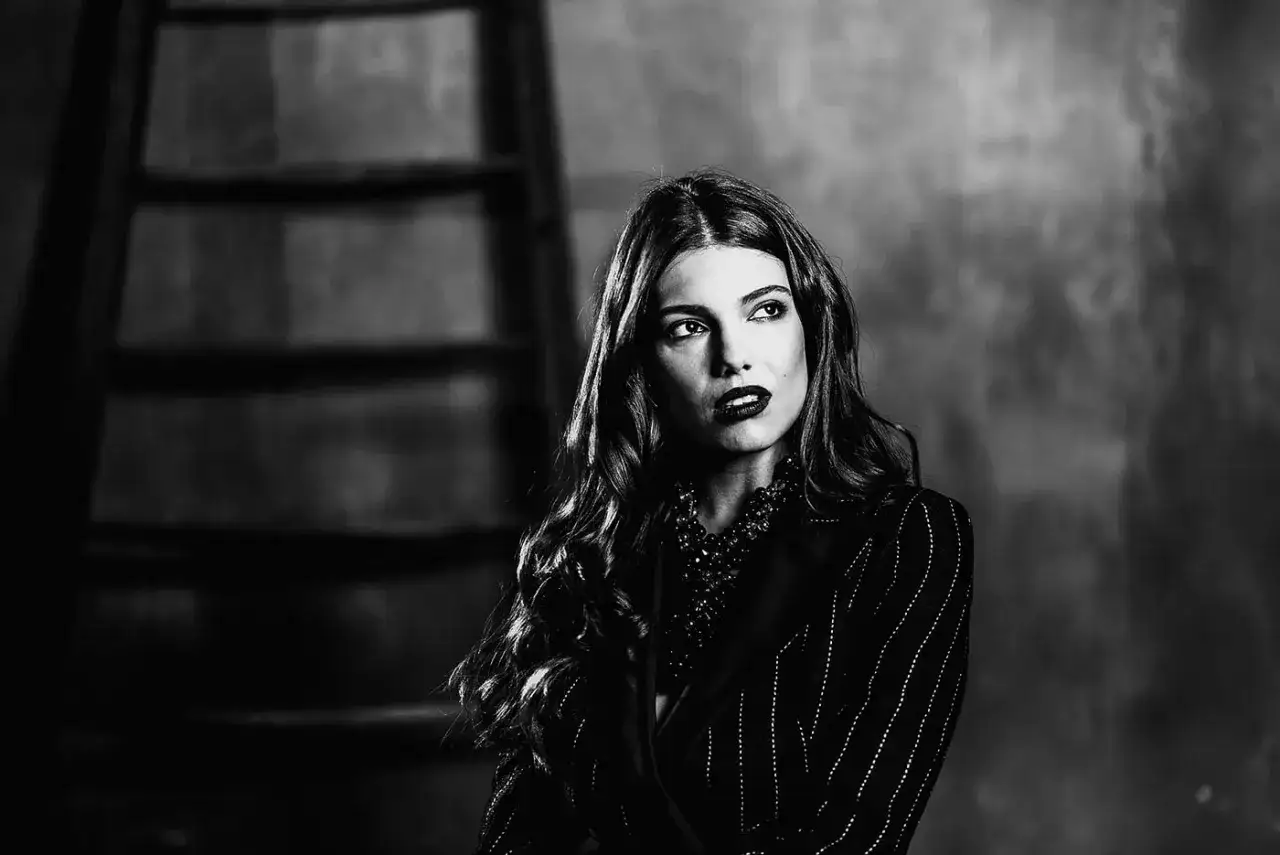 Particolari immagini bianco e nero: una donna con capelli mossi e trucco audace guarda di lato, indossando una giacca a righe e una collana vistosa.