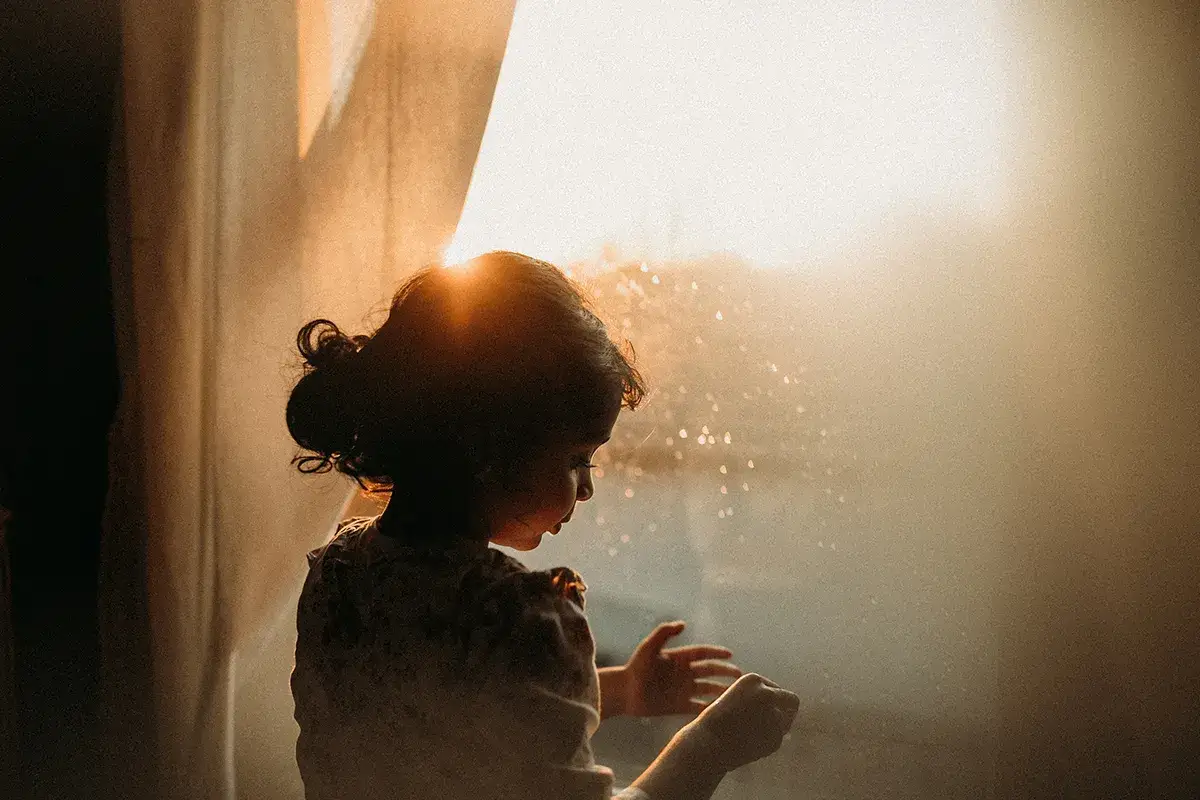 Bambina guarda fuori dalla finestra, illuminata dal sole del tramonto. Le ombre immagini creano un'atmosfera magica e sognante.