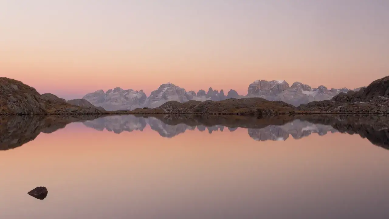 Alba sulle Dolomiti, un lago alpino riflette le cime innevate e il cielo rosa. Un sasso emerge dall'acqua, un punto di max rive in questo paesaggio sereno.