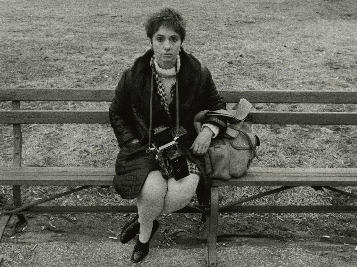 Donna con macchina fotografica vintage, seduta su una panchina, in stile Diane Arbus.