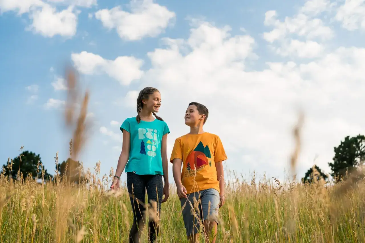 Bambini sorridono in un campo d'erba alta, con un cielo azzurro e nuvole. L'obiettivo 50 mm a cosa serve? A catturare momenti naturali come questo, con uno sfondo sfocato che fa risaltare i soggetti.