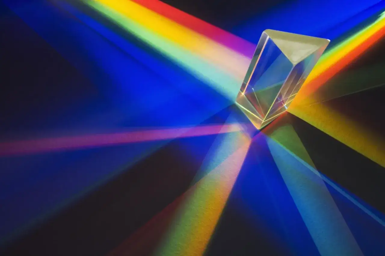 Un prisma trasparente scompone la luce, creando un arcobaleno di colori che si estende sul fondo scuro. Lo spettro del visibile si rivela in tutta la sua bellezza.