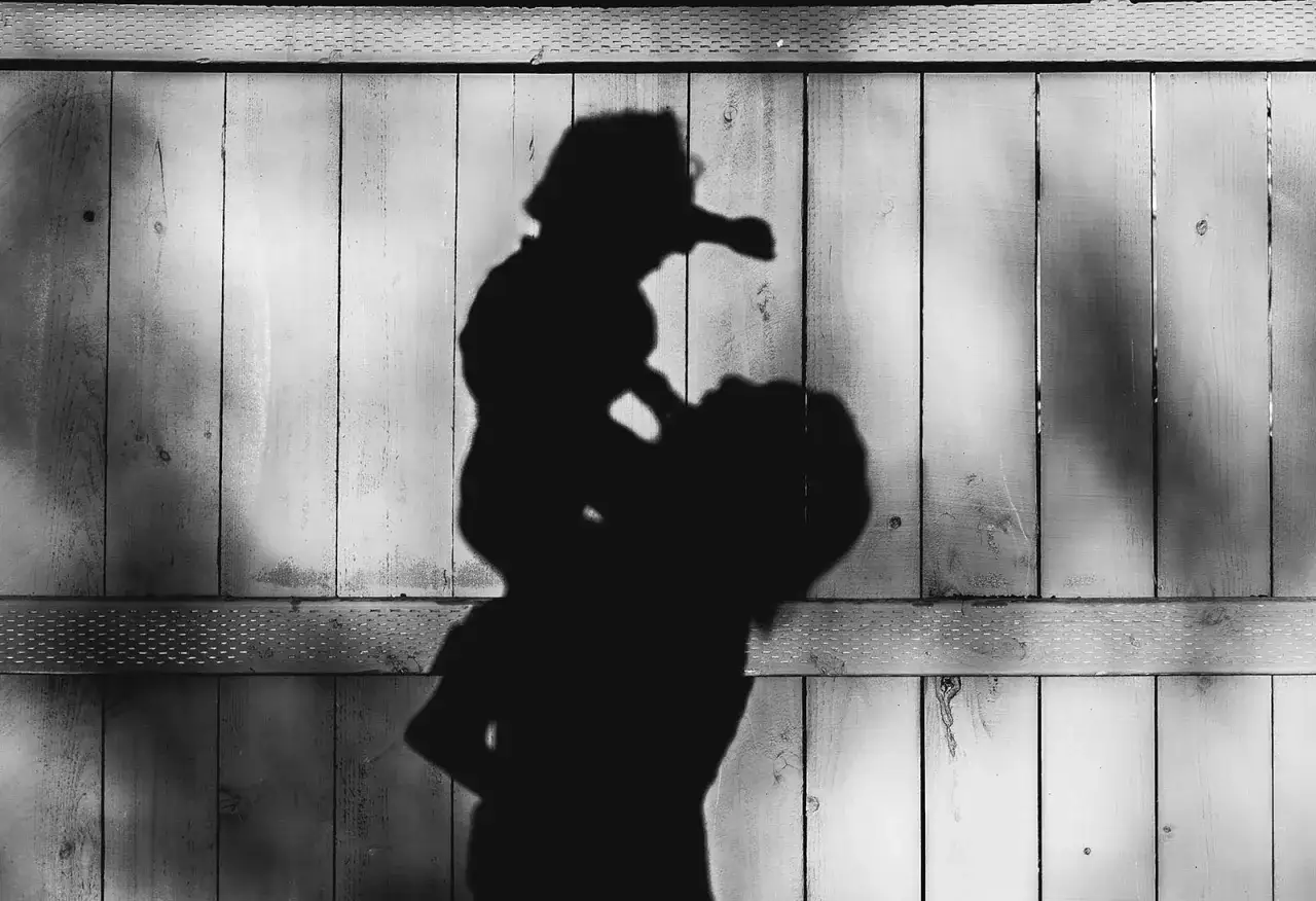 Ombra di un adulto che solleva un bambino, proiettata su una staccionata di legno sotto una luce dura.
