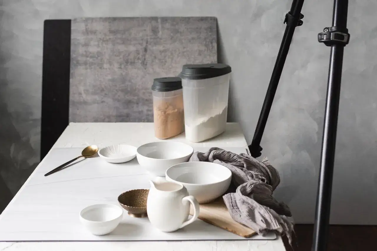 Composizione in flat lay photography: ciotole, brocca, contenitori con farina e zucchero, cucchiaio dorato, su un tavolo bianco.