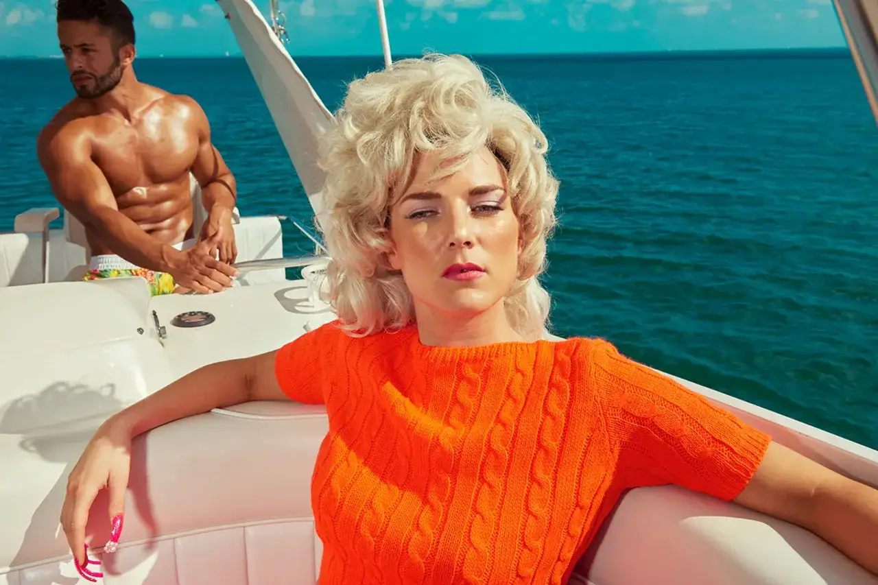 Kourtney Roy su yacht, con un uomo muscoloso sullo sfondo, mare blu e cielo limpido.