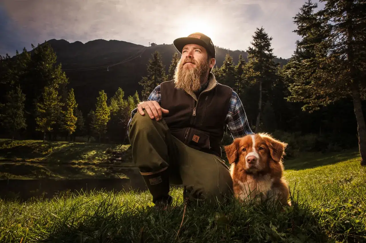Uomo con barba e cane in un bosco al tramonto. La foto flash cattura un momento di quiete tra natura e amicizia.