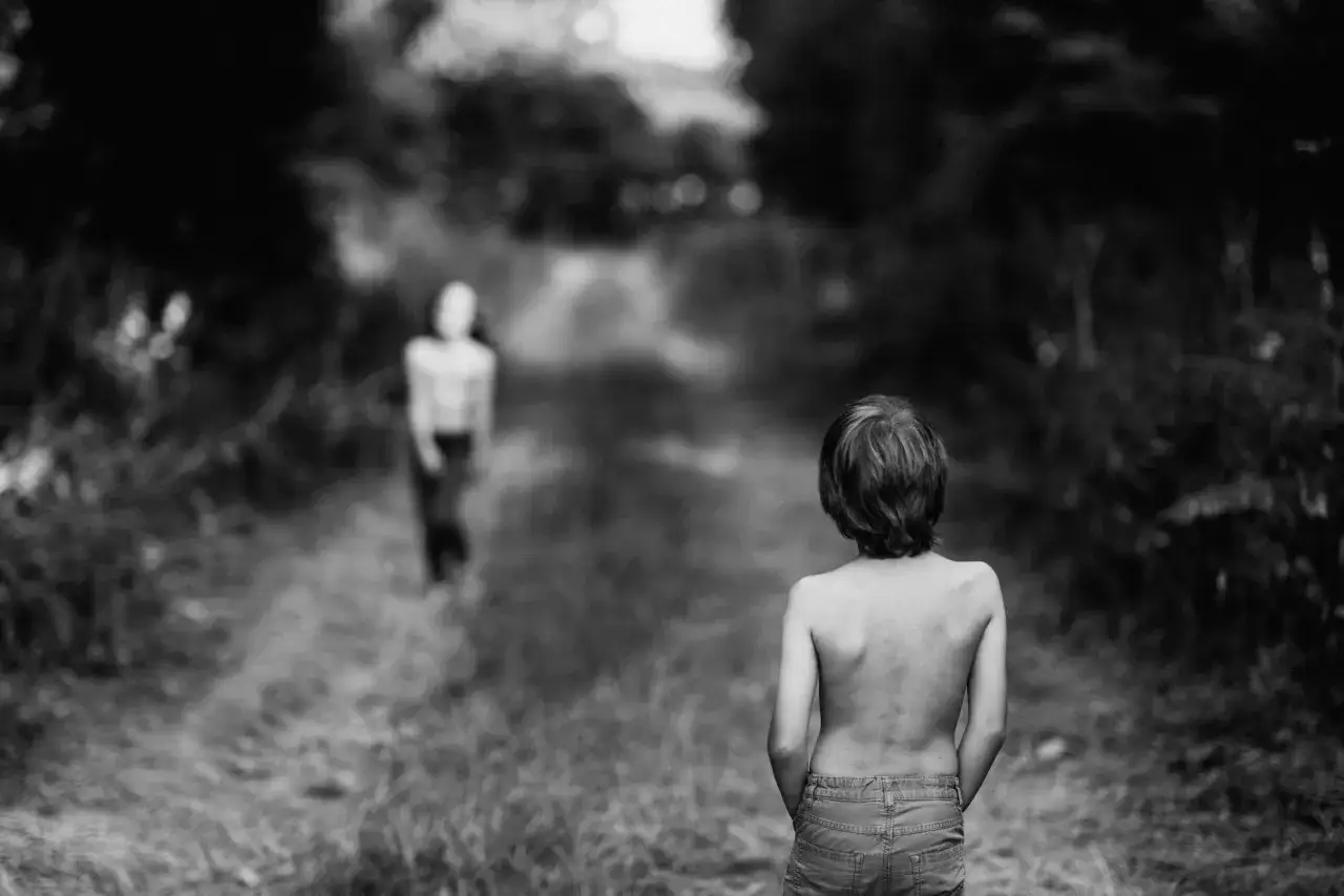 Bambino a torso nudo con le mani nelle tasche dei jeans, in un sentiero boscoso. Un'altra figura è sfocata in lontananza.