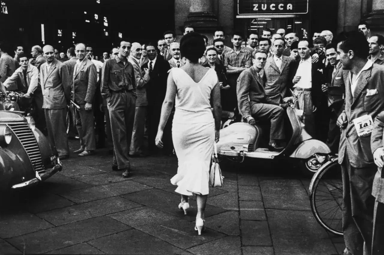 Una donna elegante cammina tra la folla, circondata da uomini e scooter, in una scena che cattura l'essenza delle foto iconiche italiane.