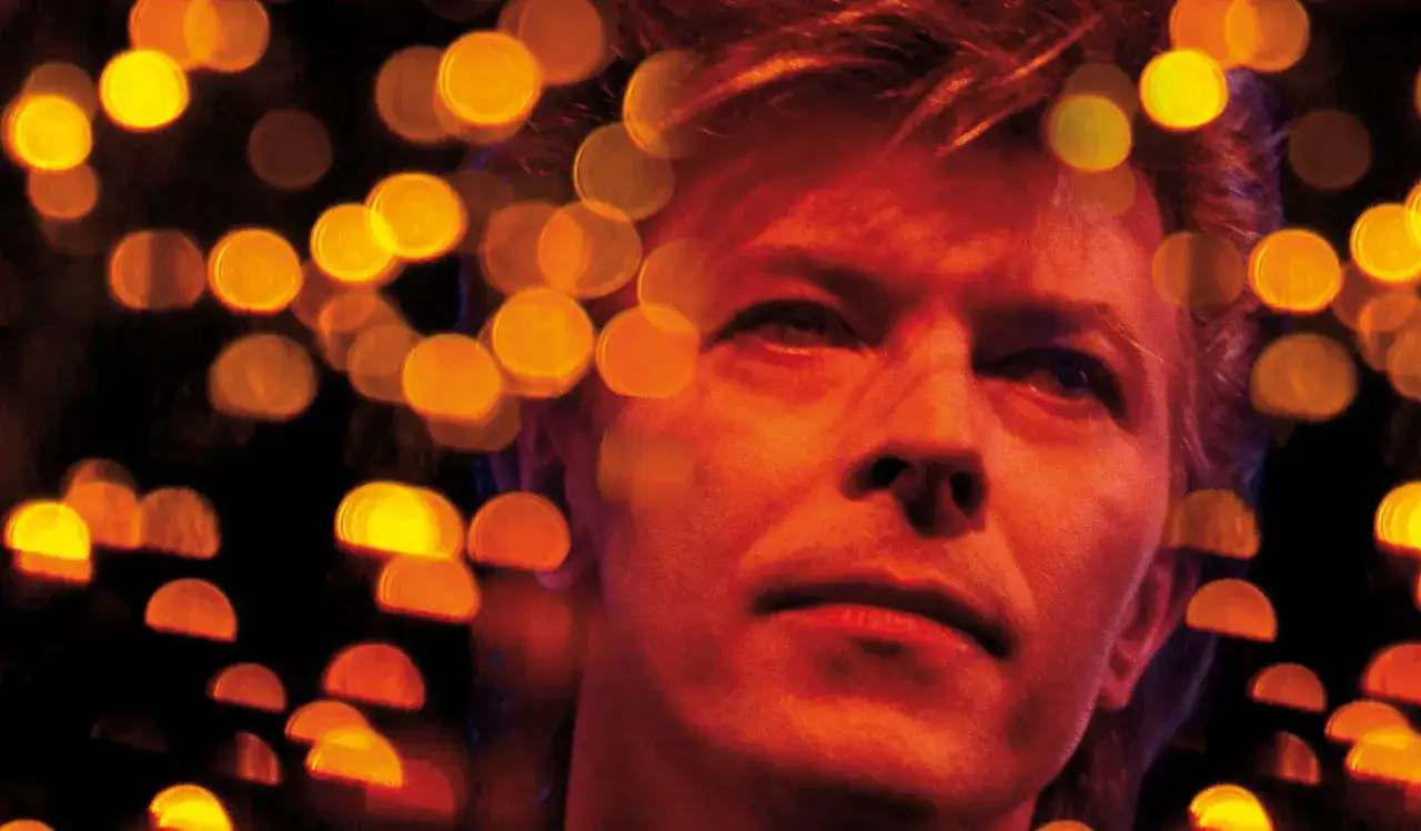 Un primo piano intenso di David Bowie, catturato in una guido harari foto, circondato da luci sfocate color oro e arancio.